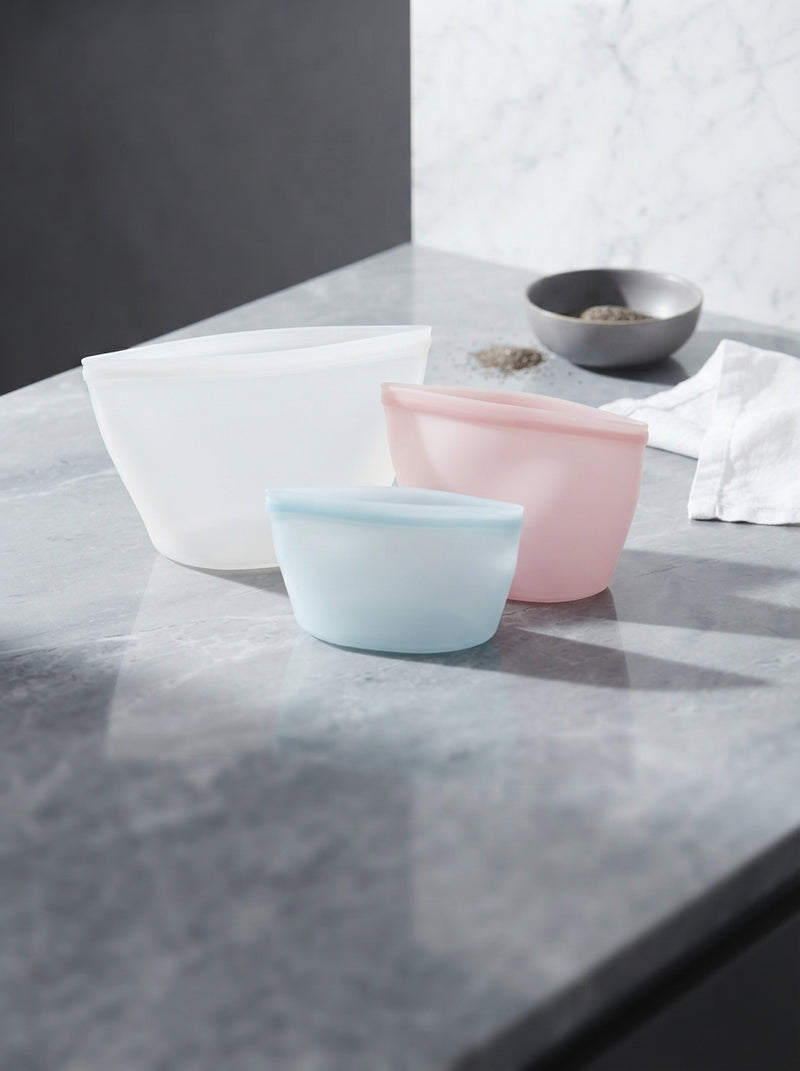 Three silicone food storage bags of varying sizes placed on a slate gray marble countertop in a bright, modern kitchen.