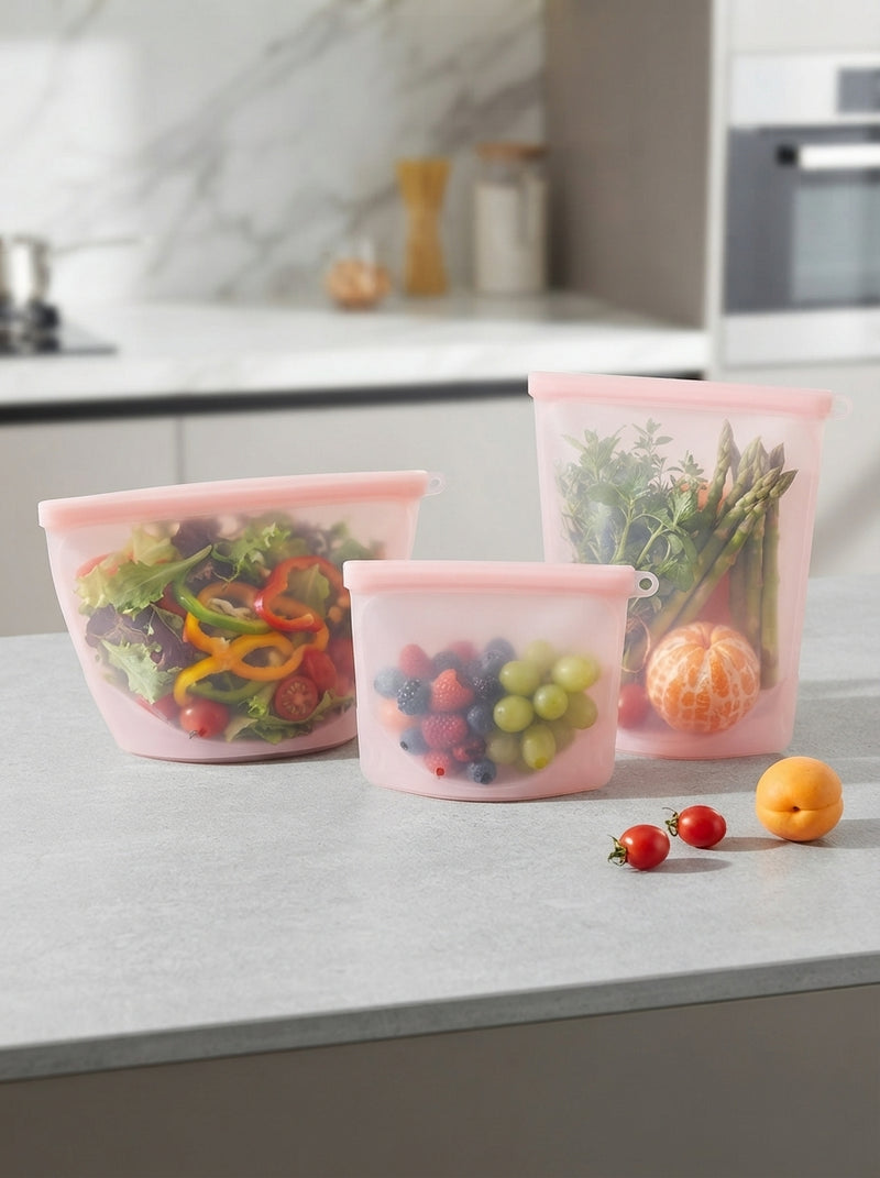 Three sizes of reusable silicone food bags filled with fresh produce arranged on a stone kitchen surface.